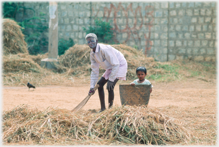 Man smiling and sweeping by hay pile, child watching.