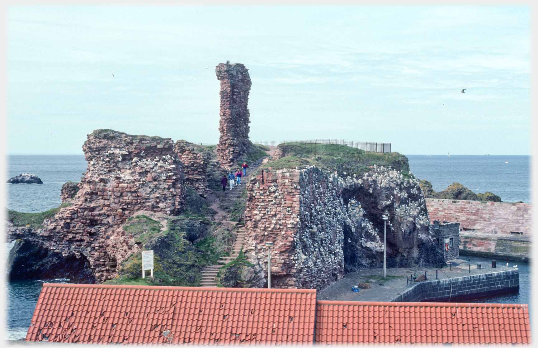 Ruins of building with ruined tower in centre, sea beyond, an roof with holes in foreground.