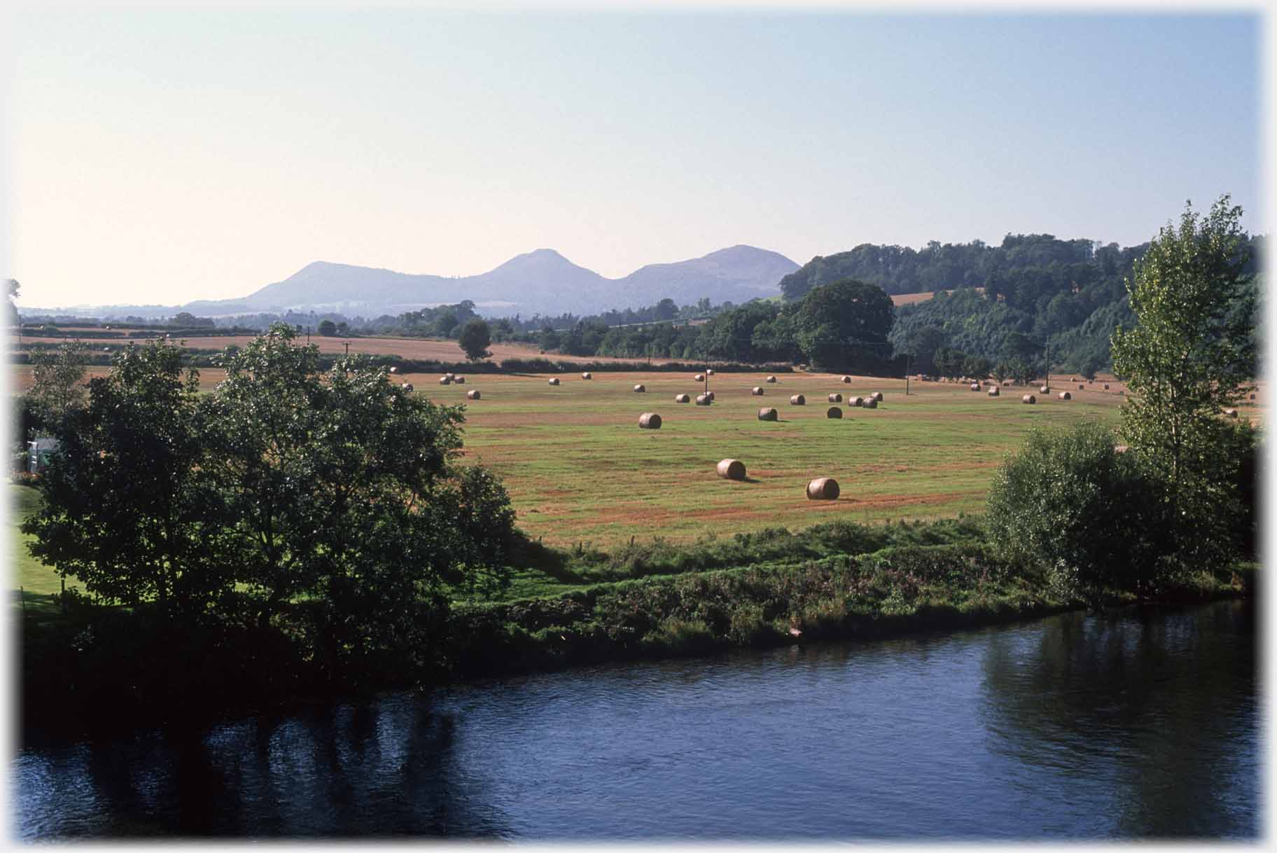 Field by river with bales and three peaked hill beyond.