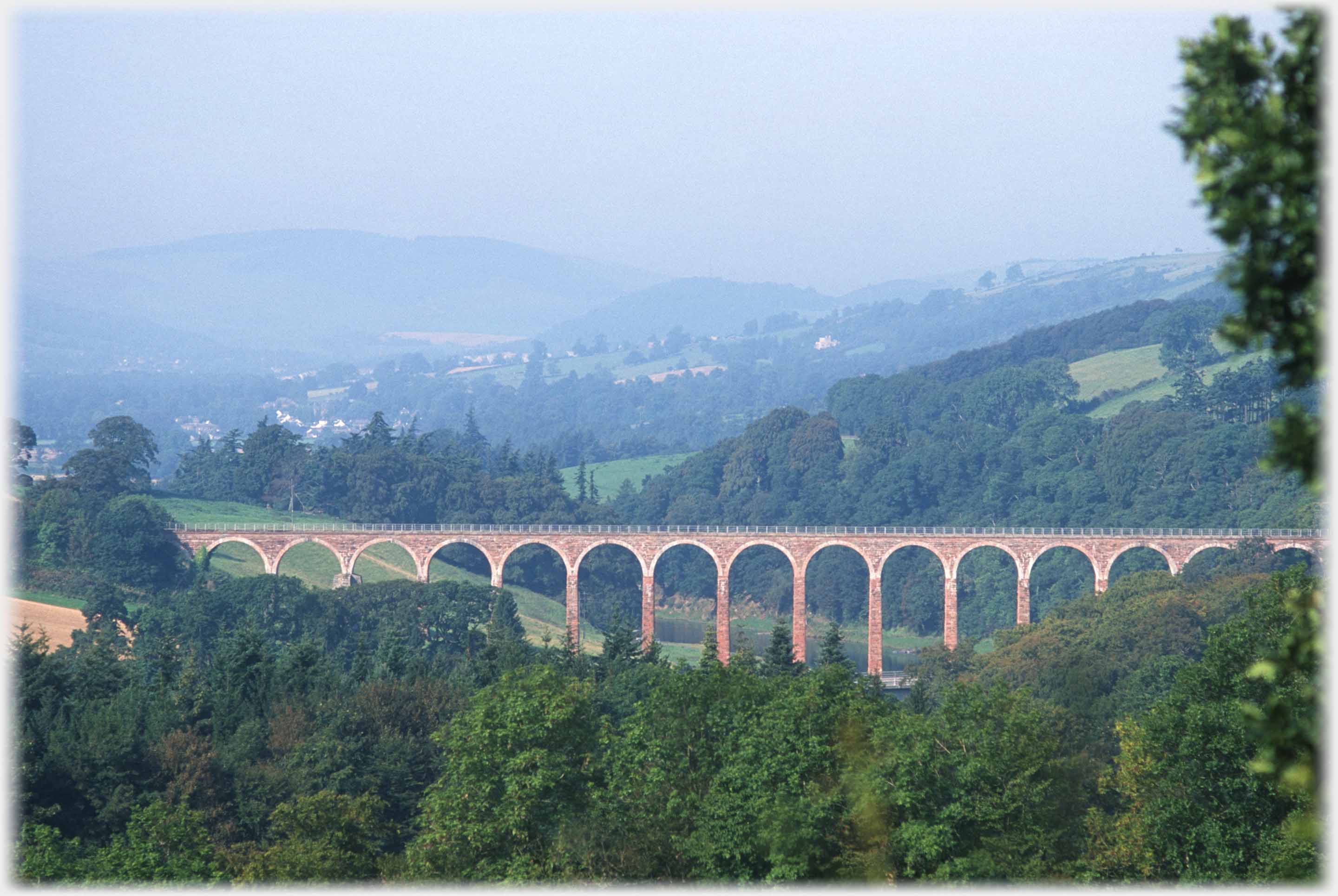 Viaduct spanning treed valley with distant houses and hills.