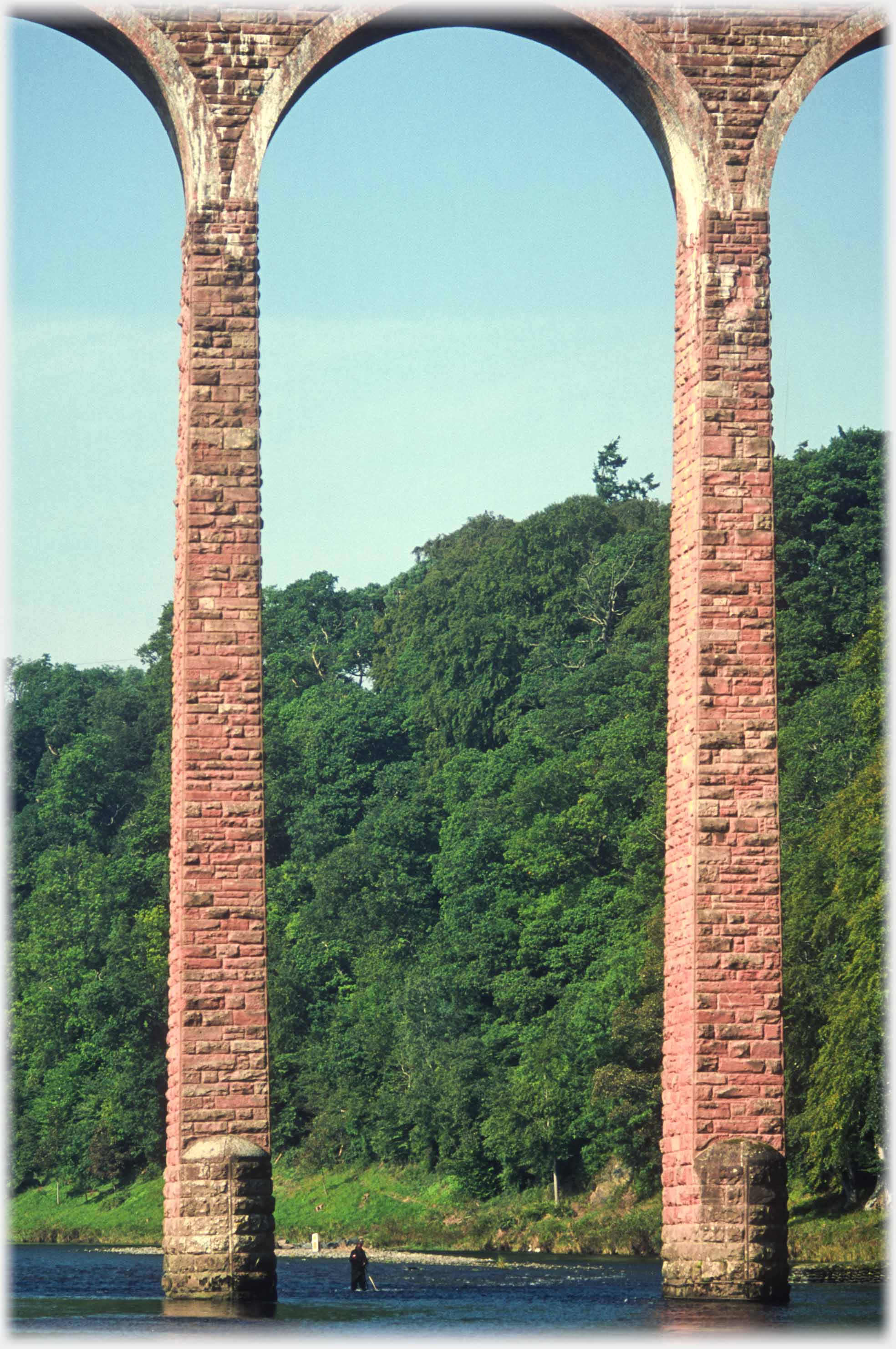 Looking up from river level at one arch of viaduct, fisherman in river.