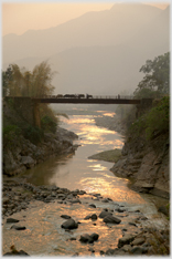 Figure and buffalo crossing bridge over sunset lit river.