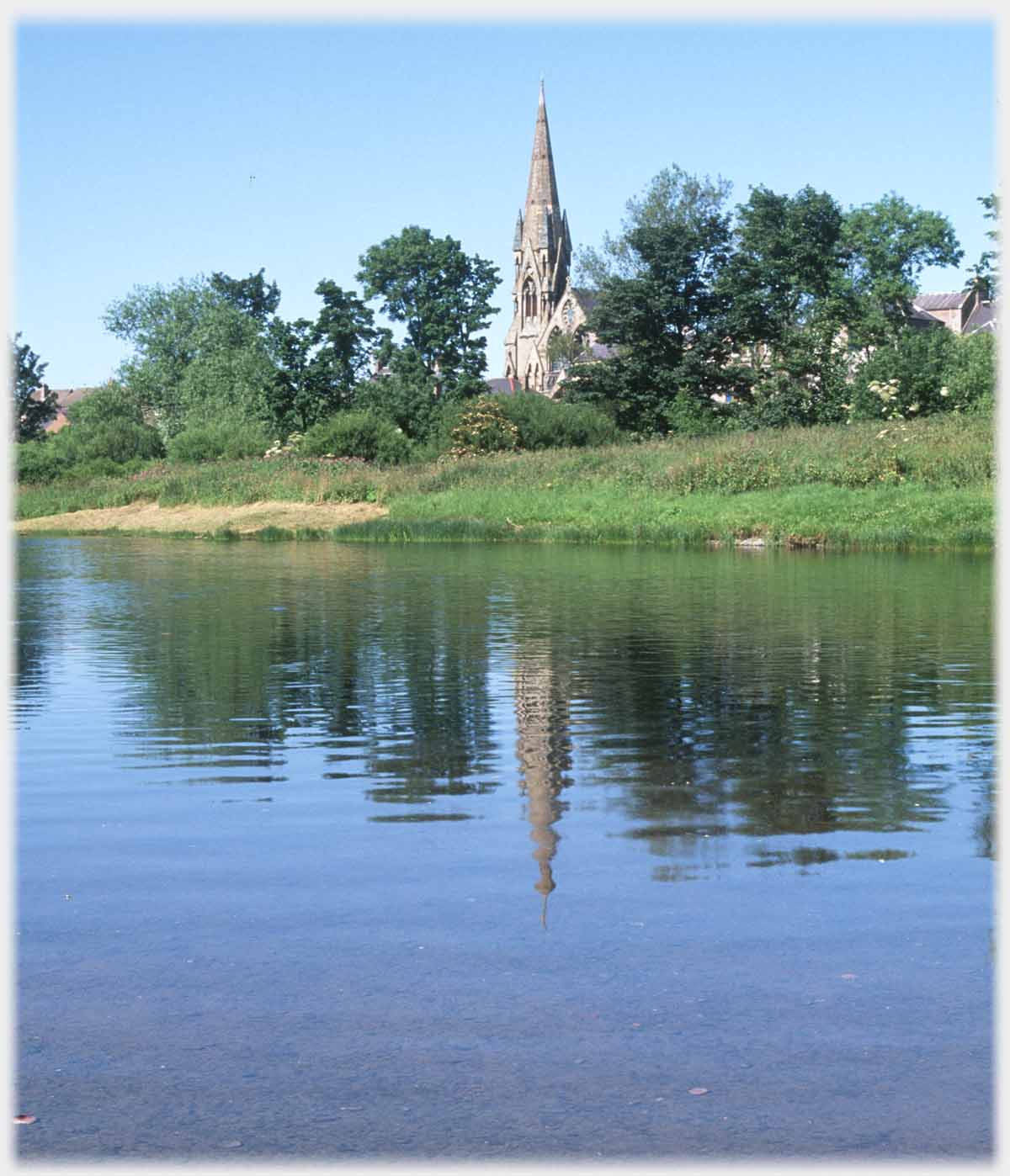 Spired church between trees reflected in river.