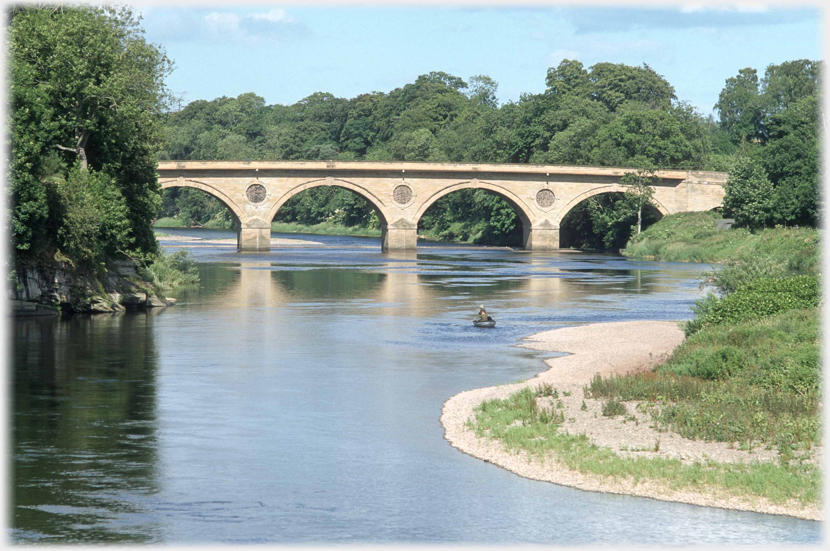 Four arches of bridge between trees, boat on river.