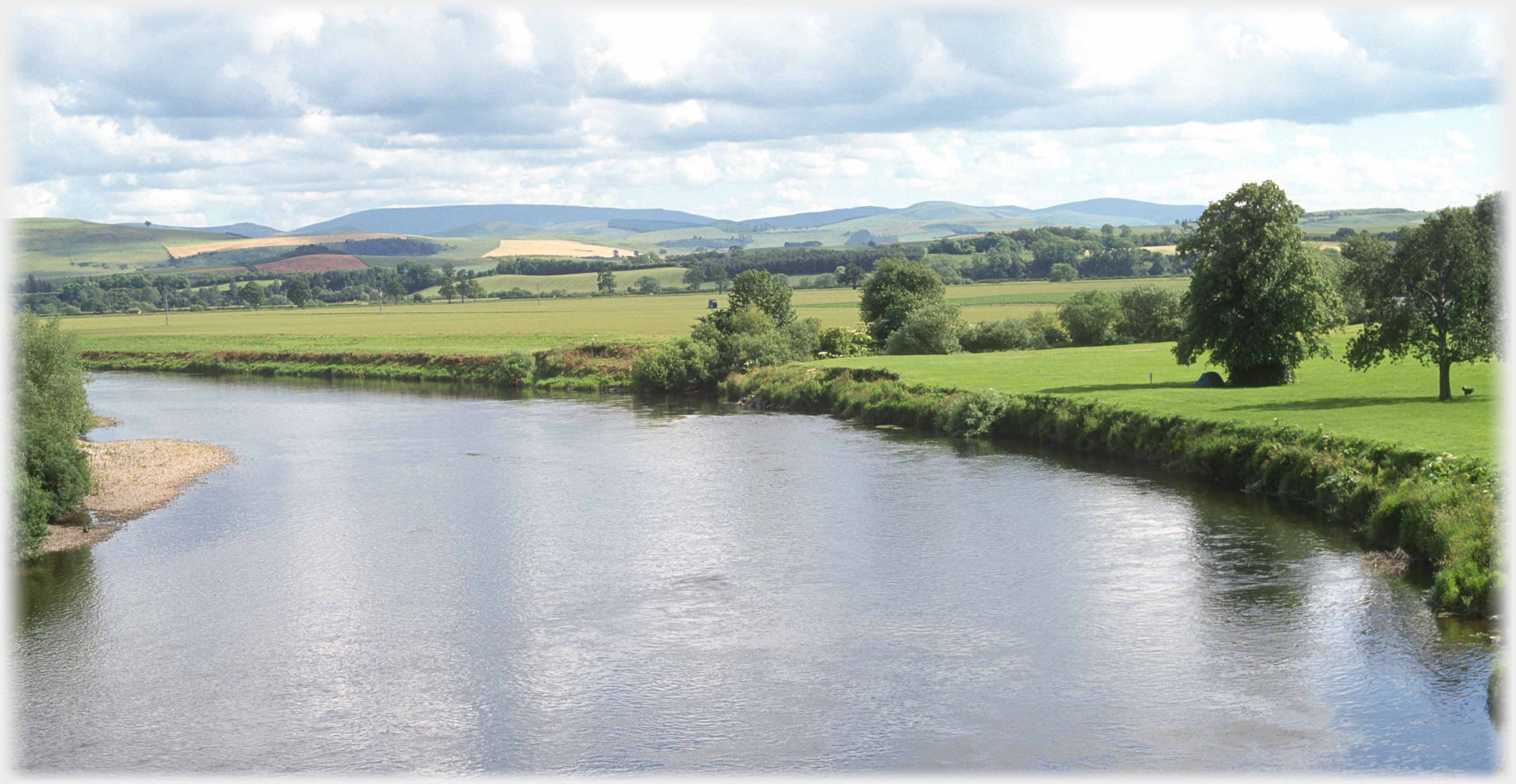 River in foreground, landscape of trees and fields leading towards hills.