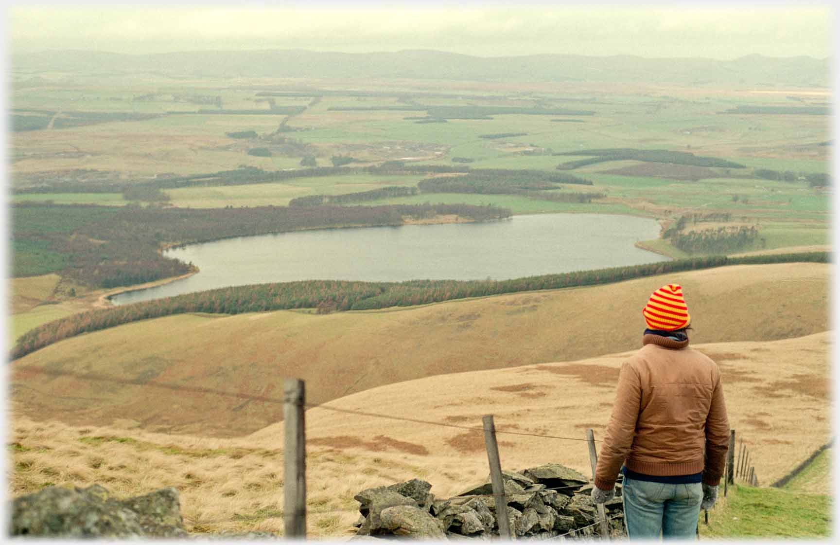 Man in woolly hat and gloves looking out at loch and plane beyond.