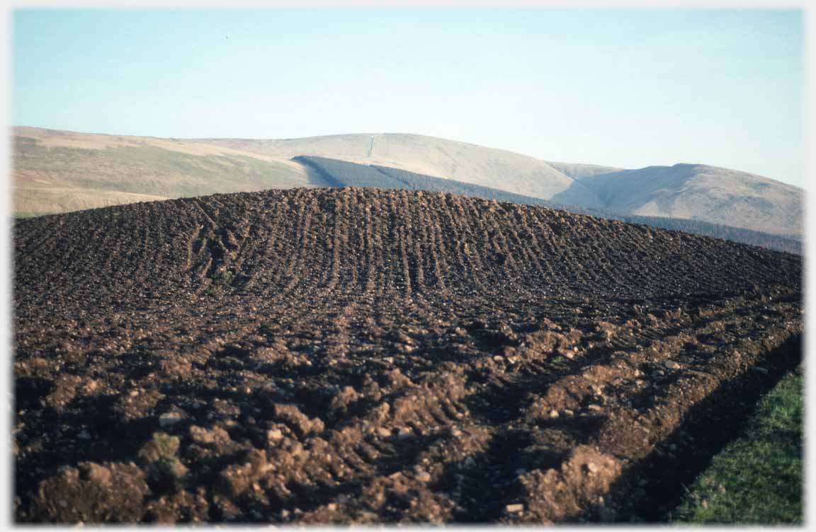 Dark furrowed soil on knoll with hills beyond.