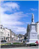 Statue on plinth in open area by road and shops.