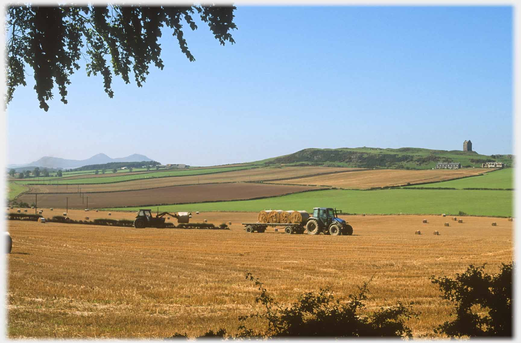 Two tractors loading bales with tower house distant.