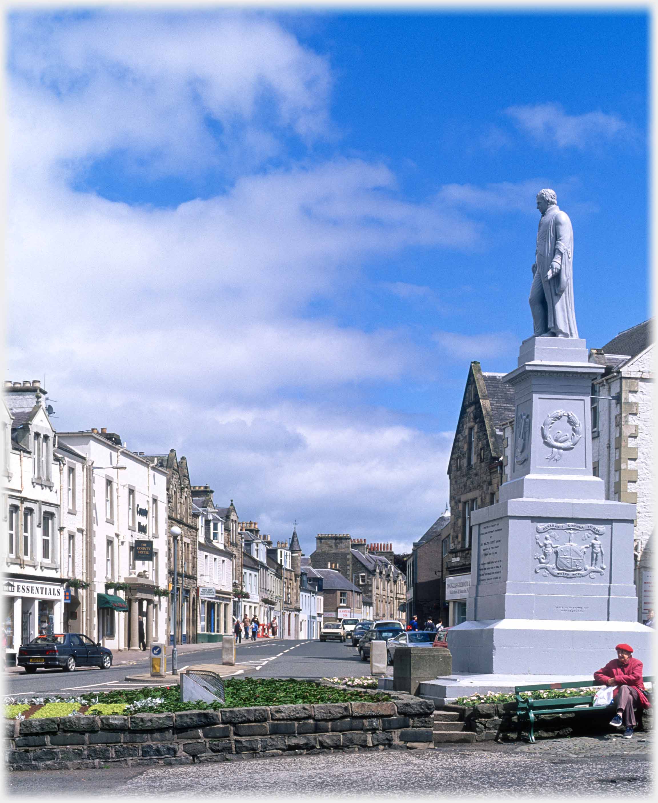 Statue on plinth by raised bed with main street behind.