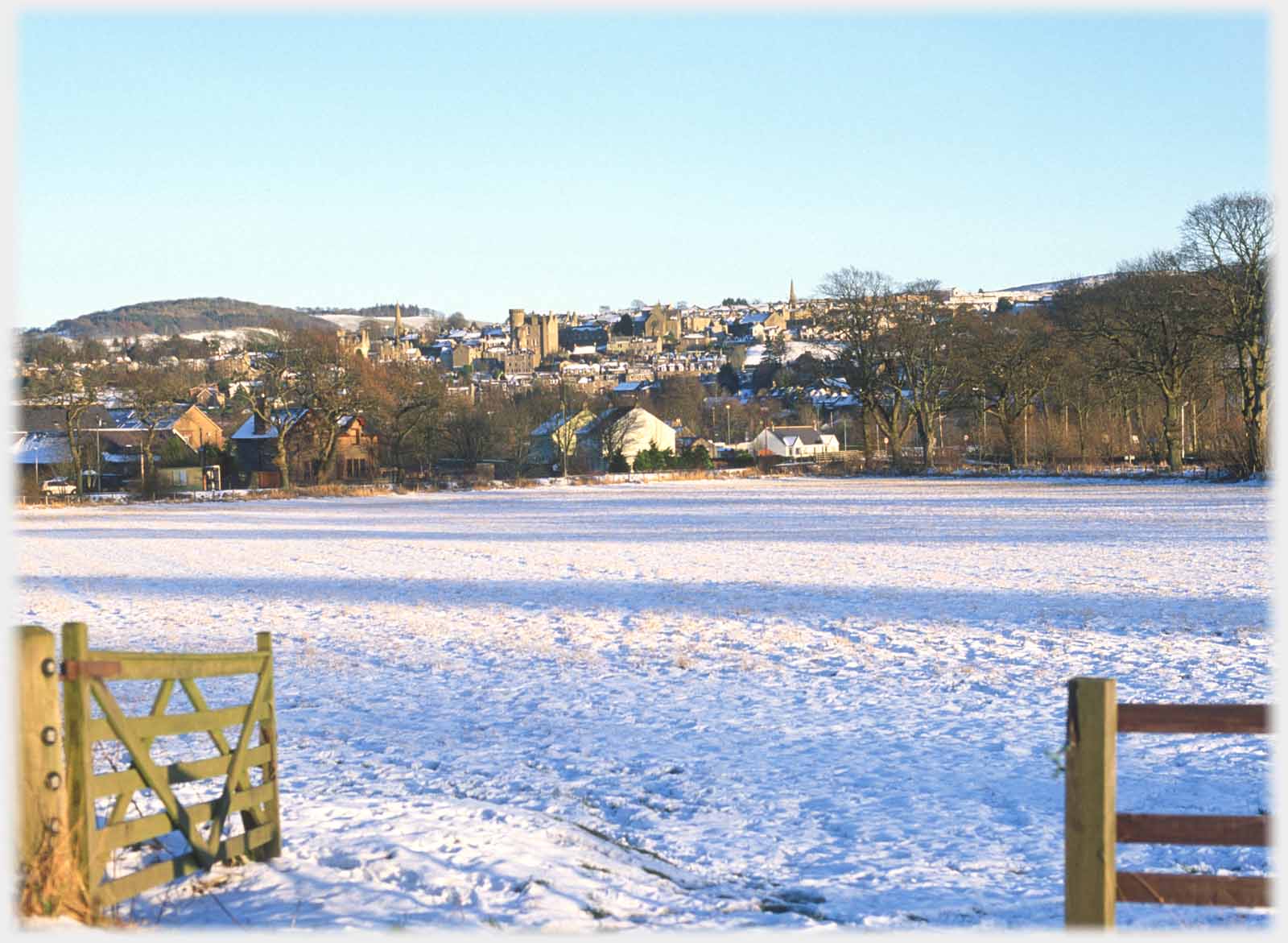 Gate open to snow covered field, town on hillside beyond.