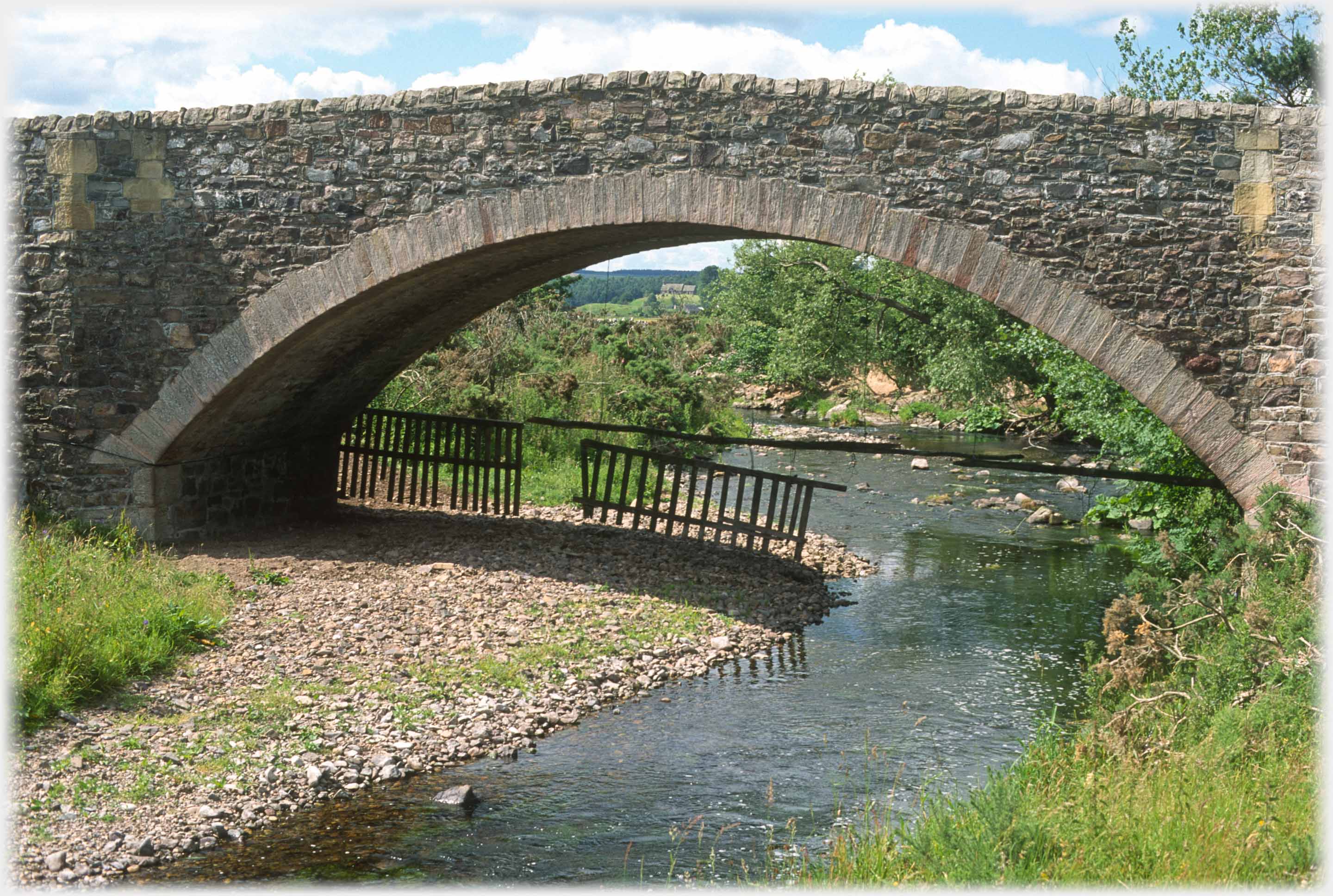 Stone arched bridge across small river, sheep fence under it.