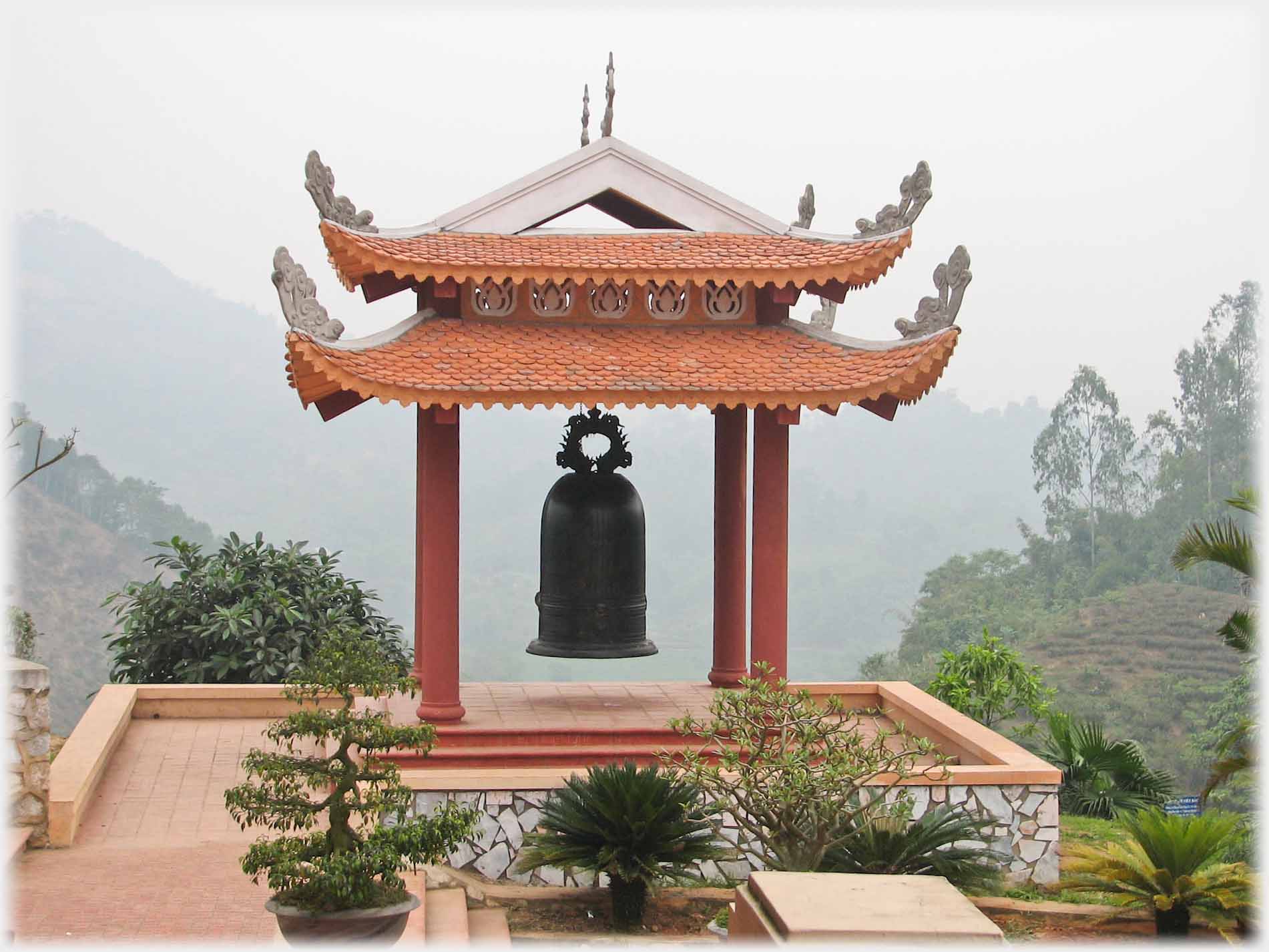 Very large bell hanging in small four pillard building,valleys and mountains beyond.