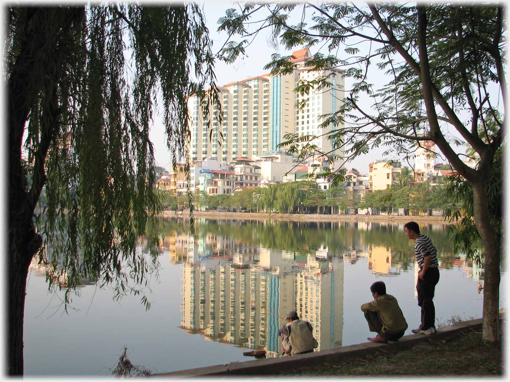 Large building reflected in lake with three fishermen on bank.