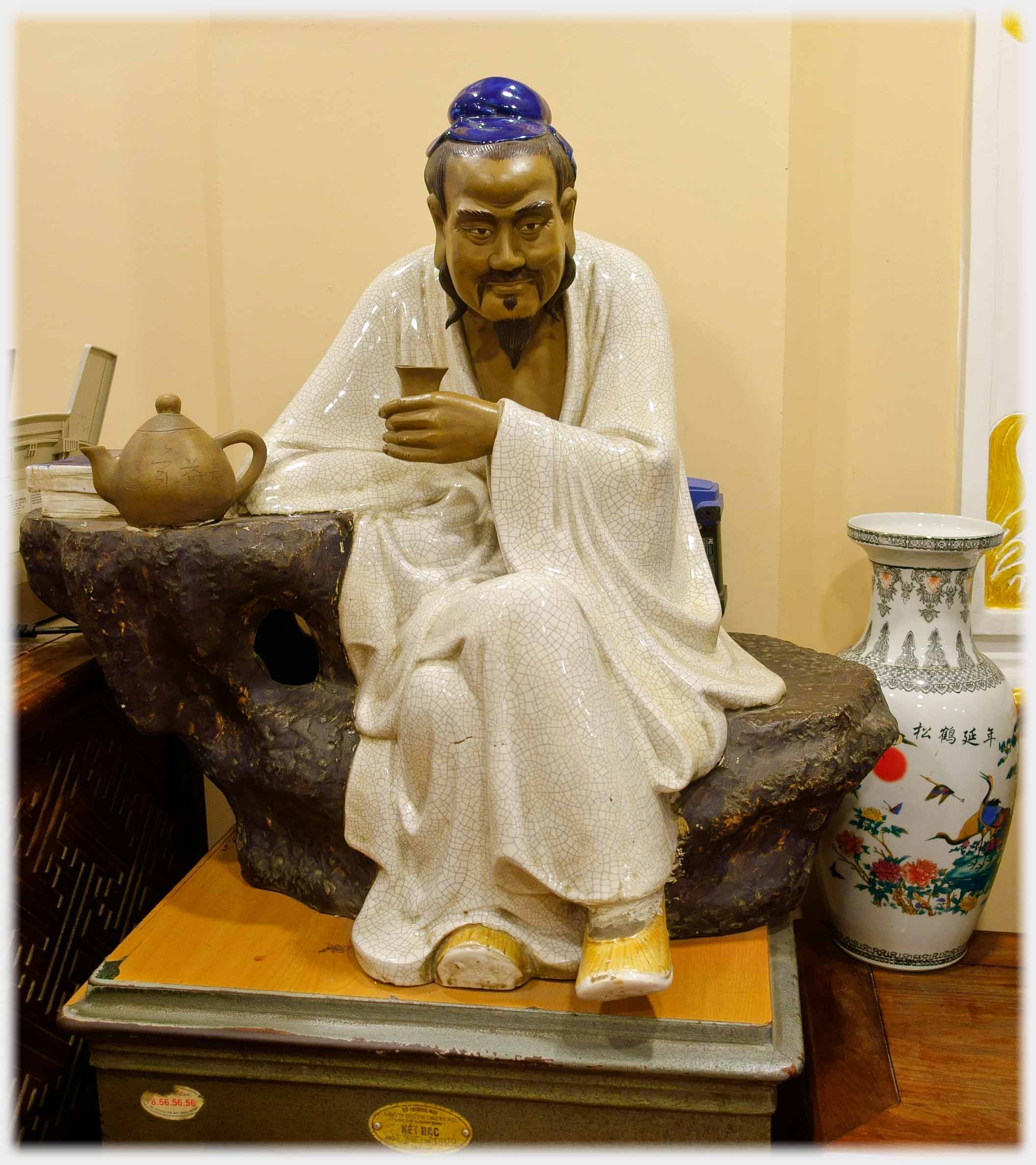 Figure in white cloak sitting with teacup by teapot.