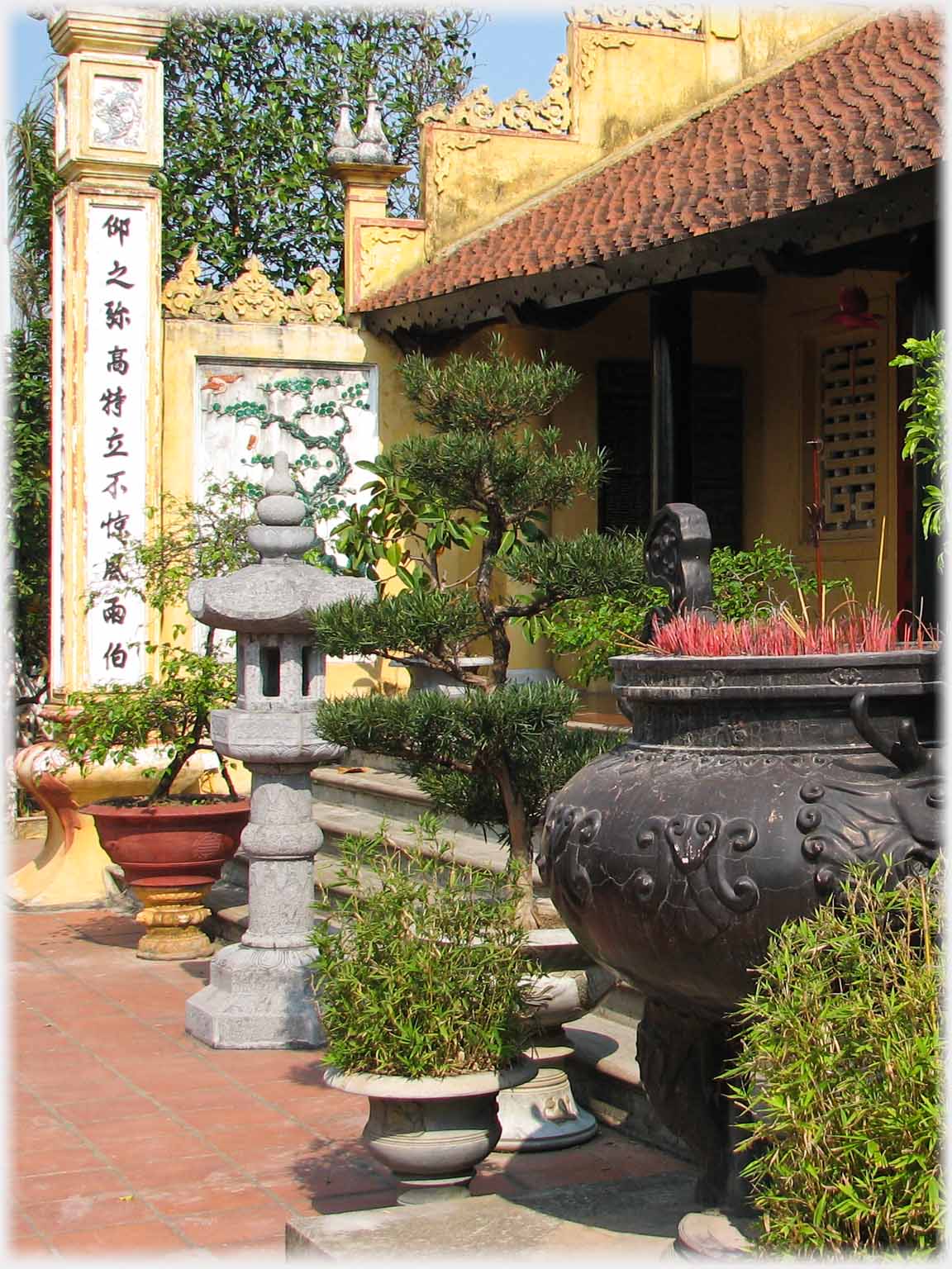 Grey stone lantern,  many potted plants and large cauldron with remains of incense sticks.