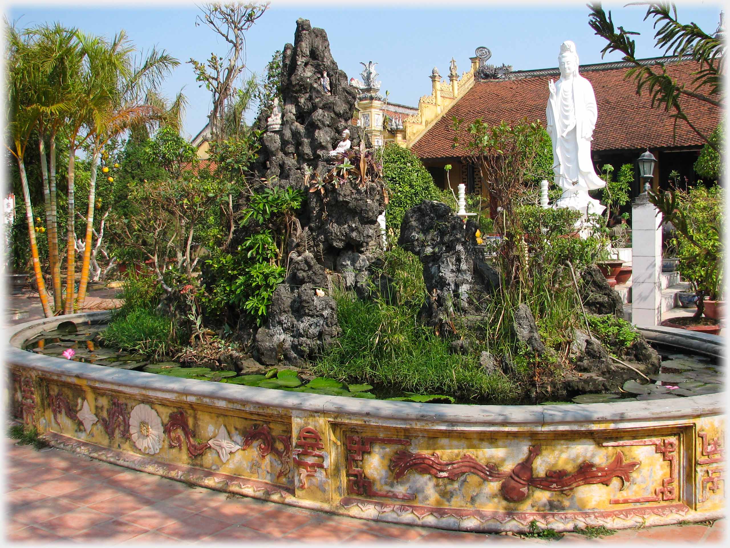 Raised pool with large rough rocks, many plants and objects on and around, white standing Buddha behind.