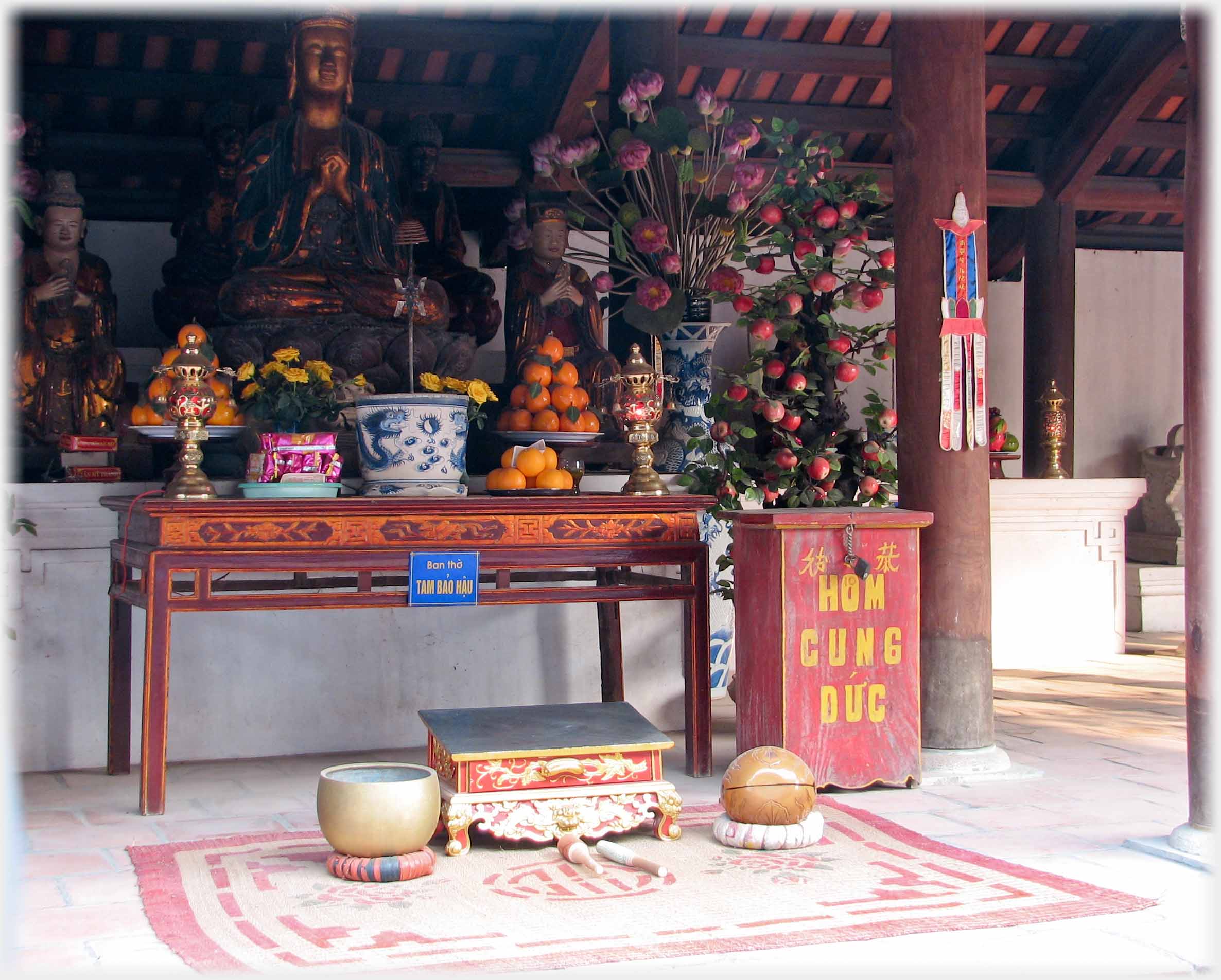 Plain table altar with gilded Buddha, fruit offerings and large red box to side.