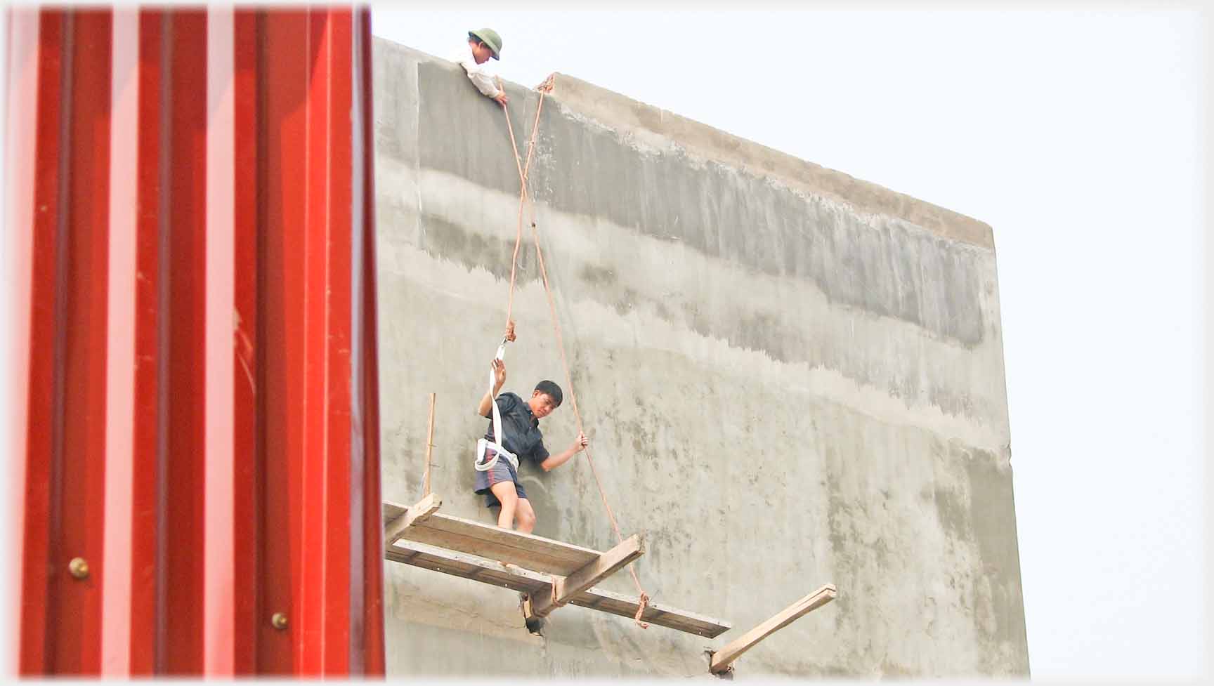 Plank walkway on upper floor, man on rope, held by another man, standing on it.