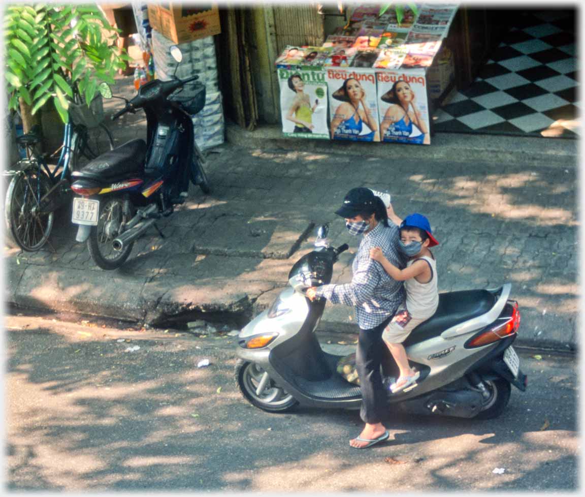 Woman on motorbike with small child behind reaching up to hold her shoulders.