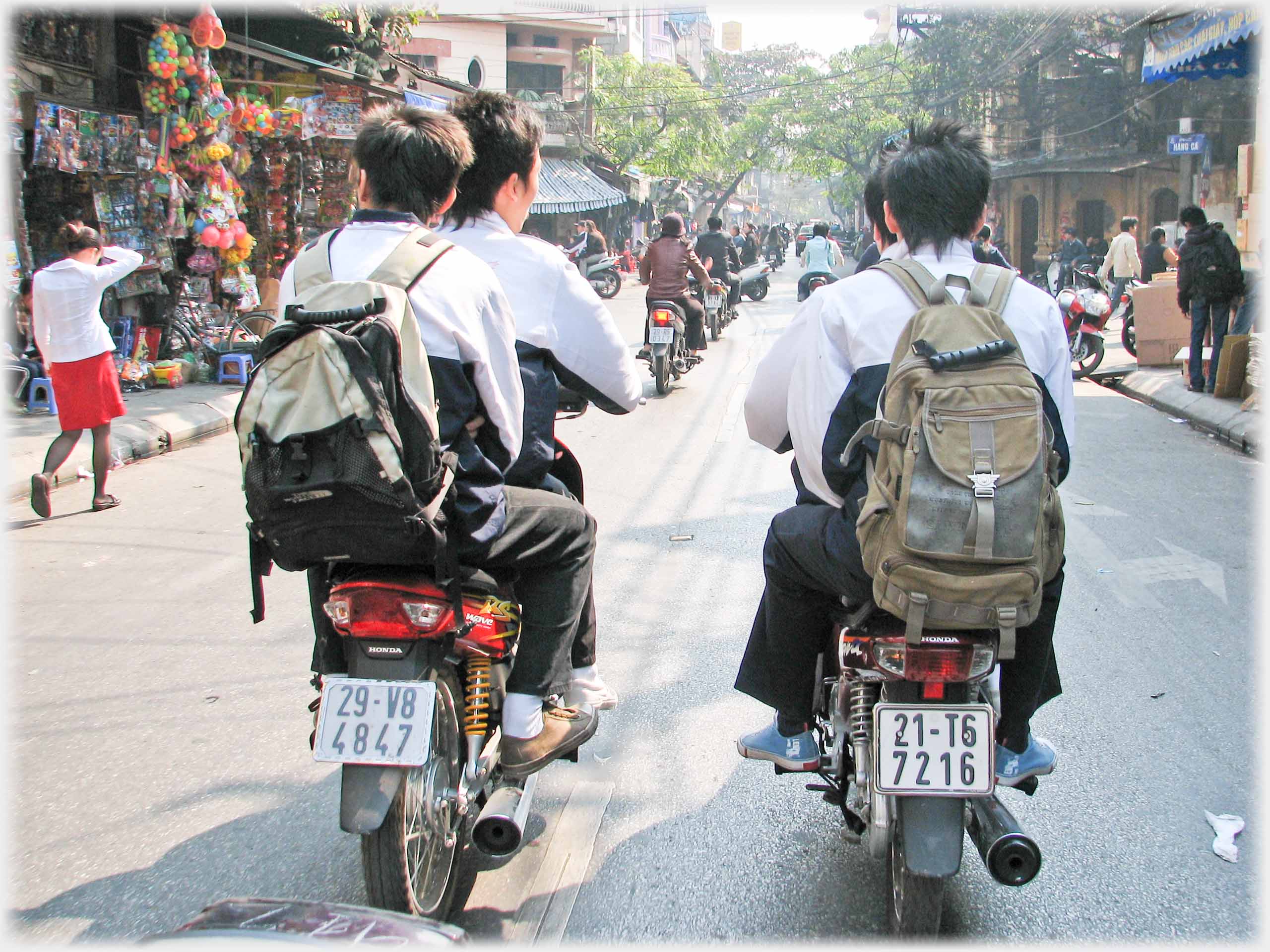 Backs of two motorbikes each with two boys in school uniform riding together.