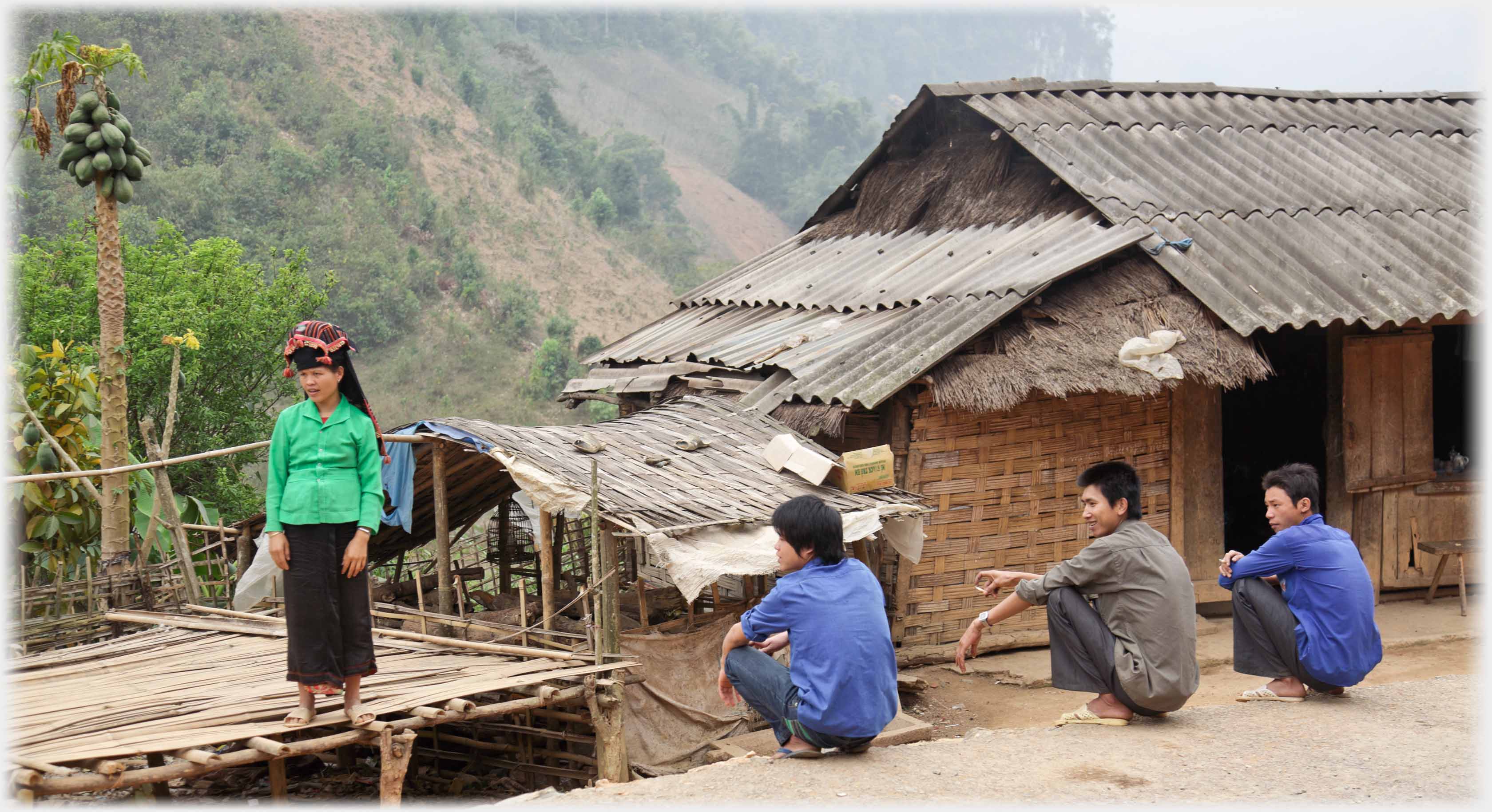 Three men squatting at road-side with cigarettes beside house and young woman standing on platform.