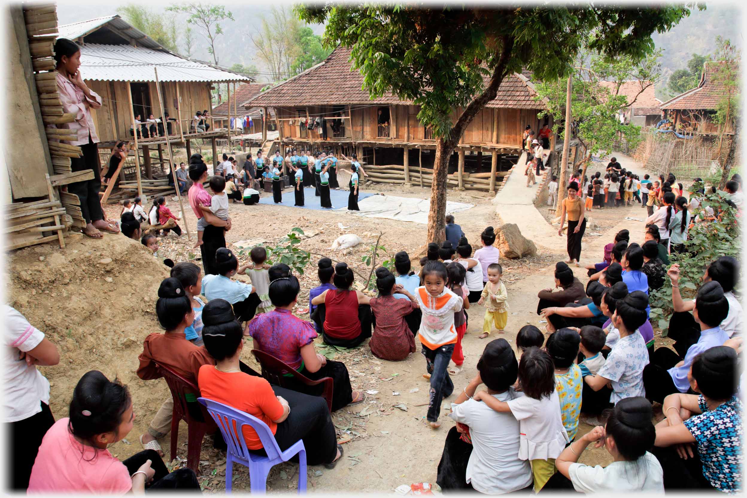 On sloping ground in village, looking over some dozens of people sitting towards the dancing display.