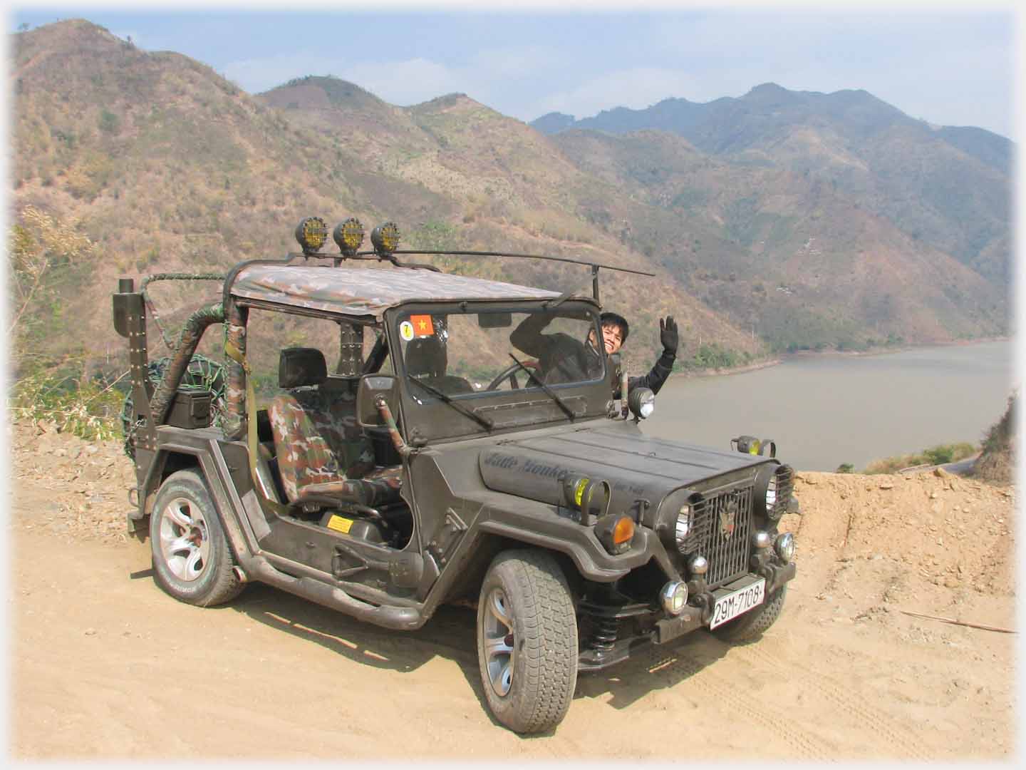 Han leaning out of Jeep waving, lake and mountains behind.