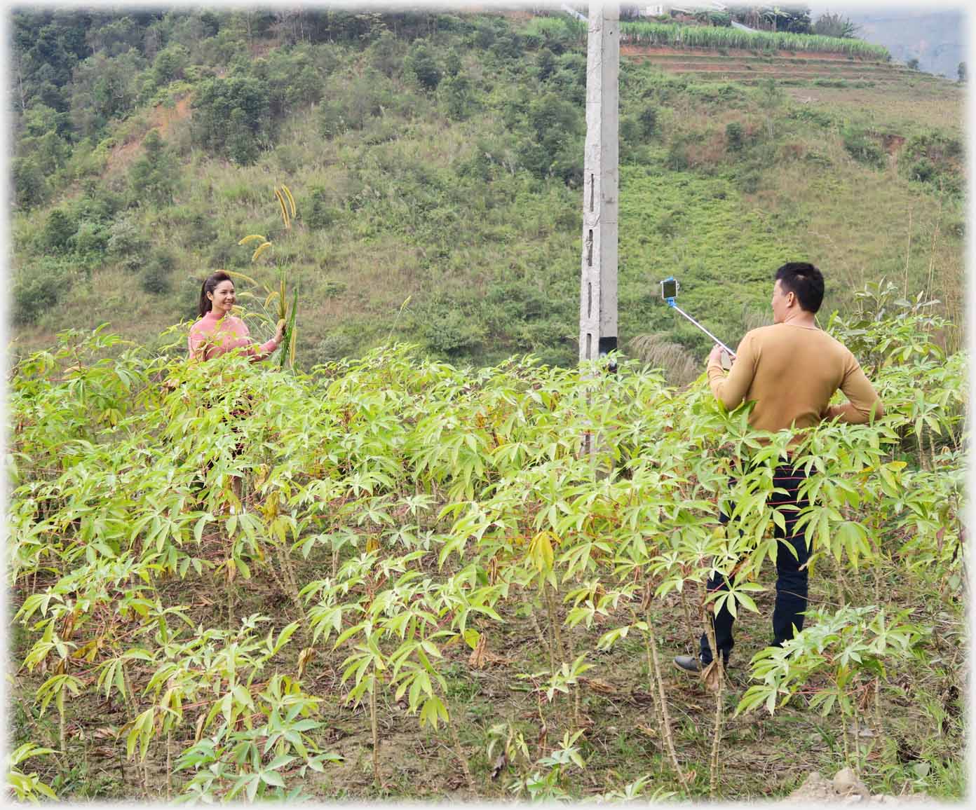 Man with phone on selfie stick in amongst plants taking photo of woman in amongst plants.