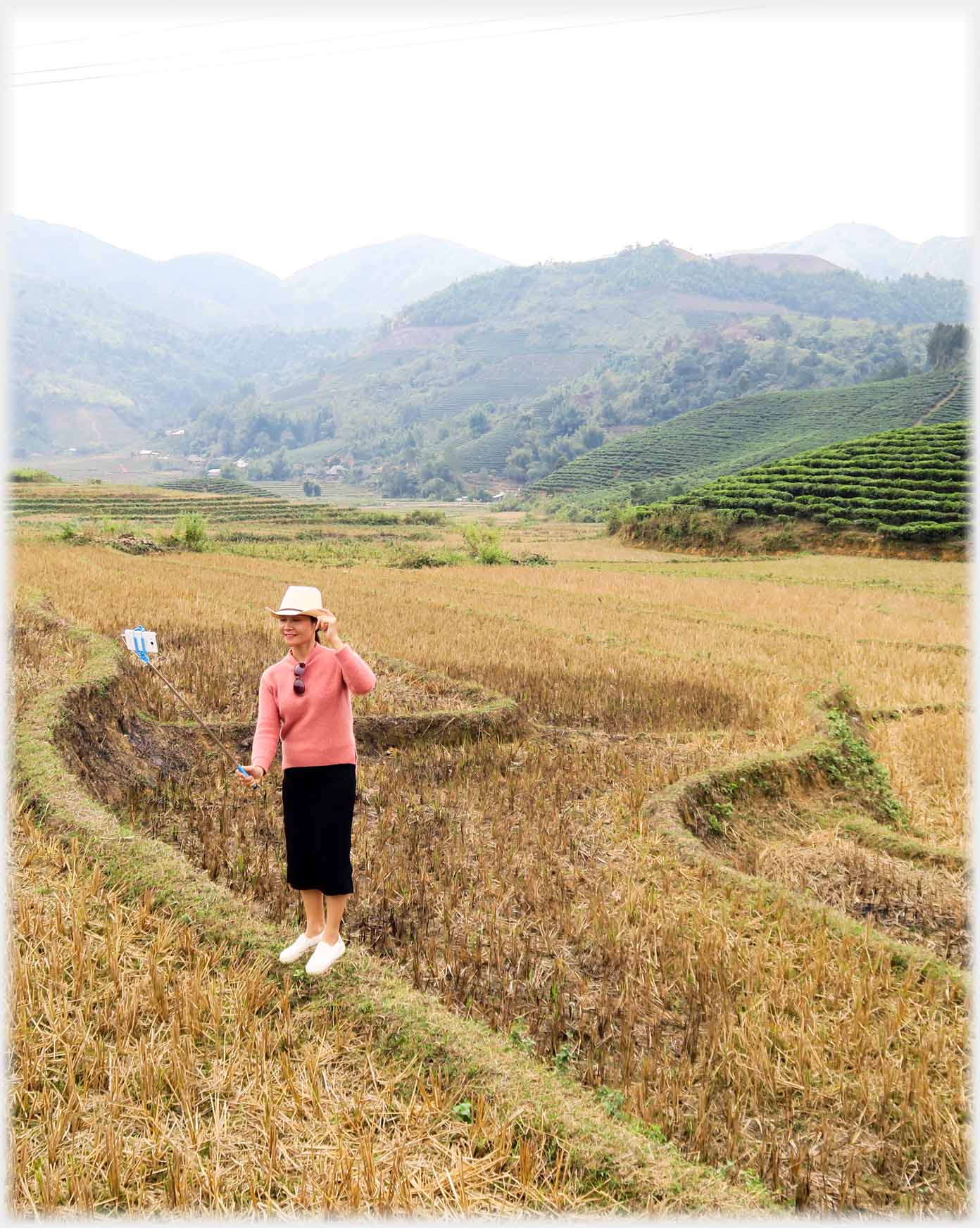 Woman standing on low narrow banking holding selfie stick with phone, country beyond.