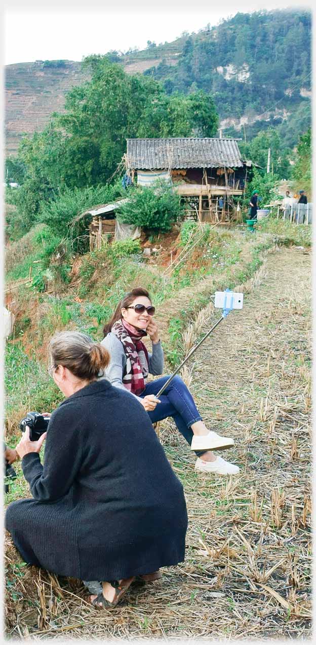 Woman sitting on bank taking picture of herself with selfie stick.