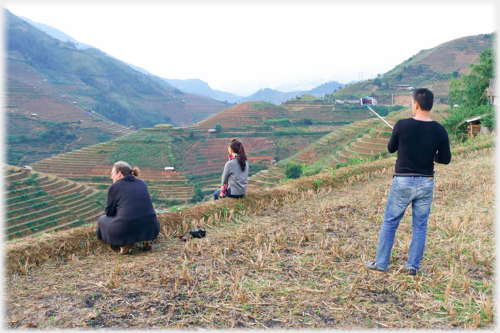 Man taking photo with phone on selfie stick of two women sitting on bank looking down valley.