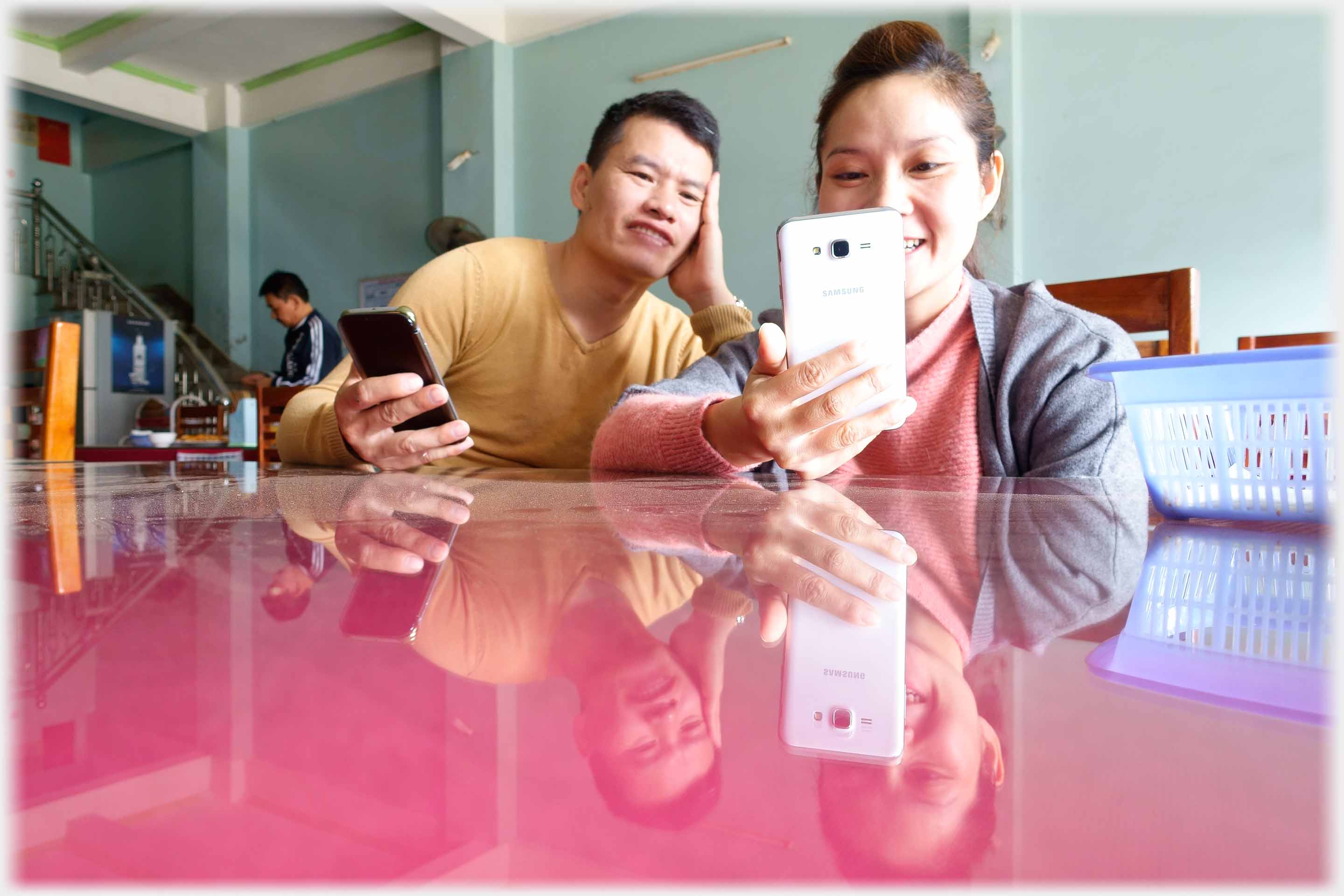 Han and Duyen both holding phones and both looking at her phone - reflected in table top.