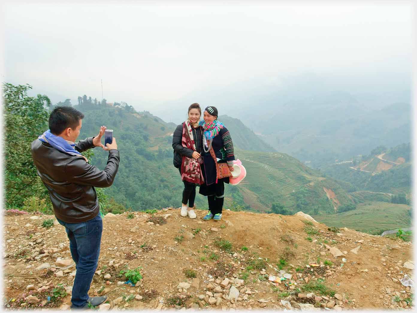 Man taking photo of two women standing together on hillock, misty hills behind.