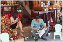 Woman sitting on step with phone, man reading paper at shop front.