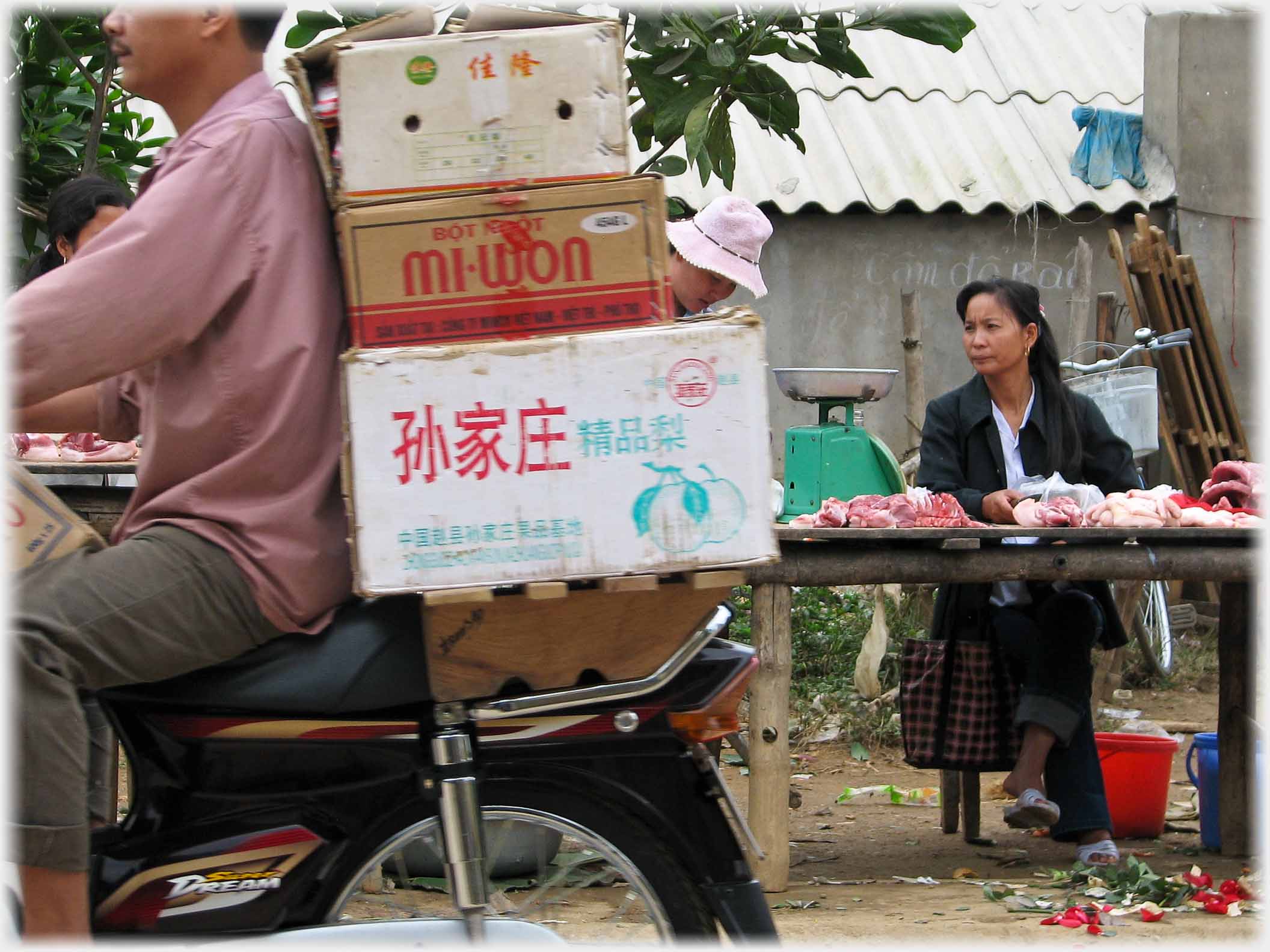 Man on mmotorbike with boxes, woman beyond behind meat stall.