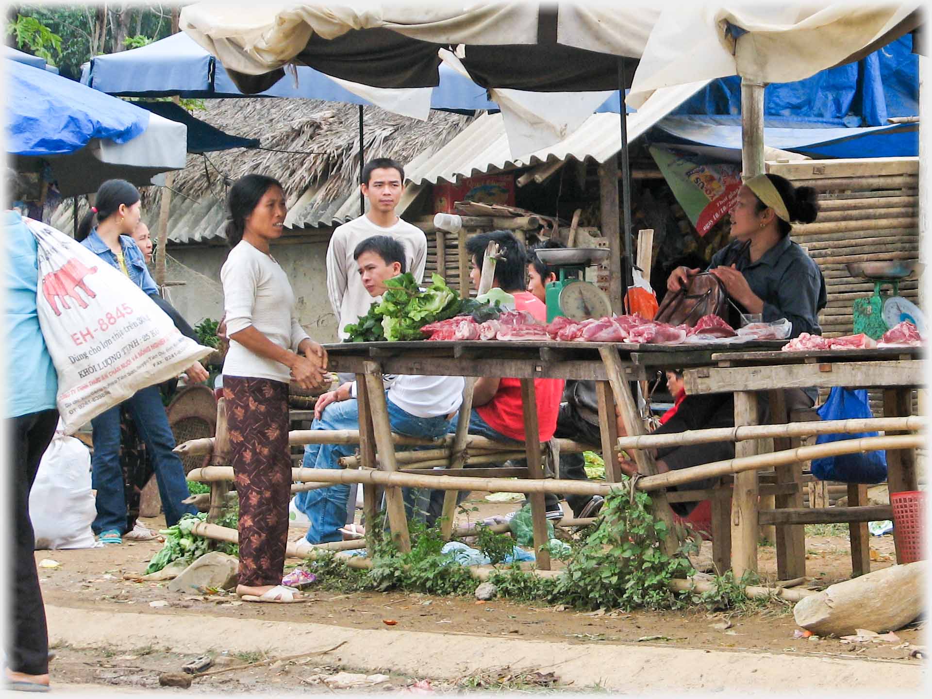 Seven people sitting and standing around meat stall.