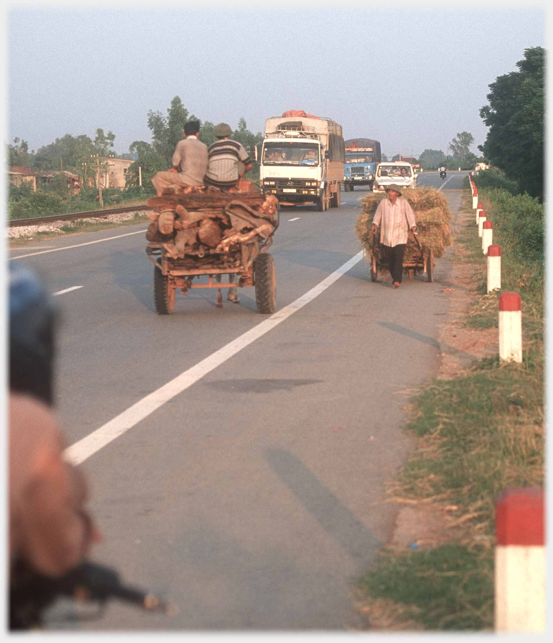 Busy road with trucks and in foreground horse and cart, and hand drawn cart.