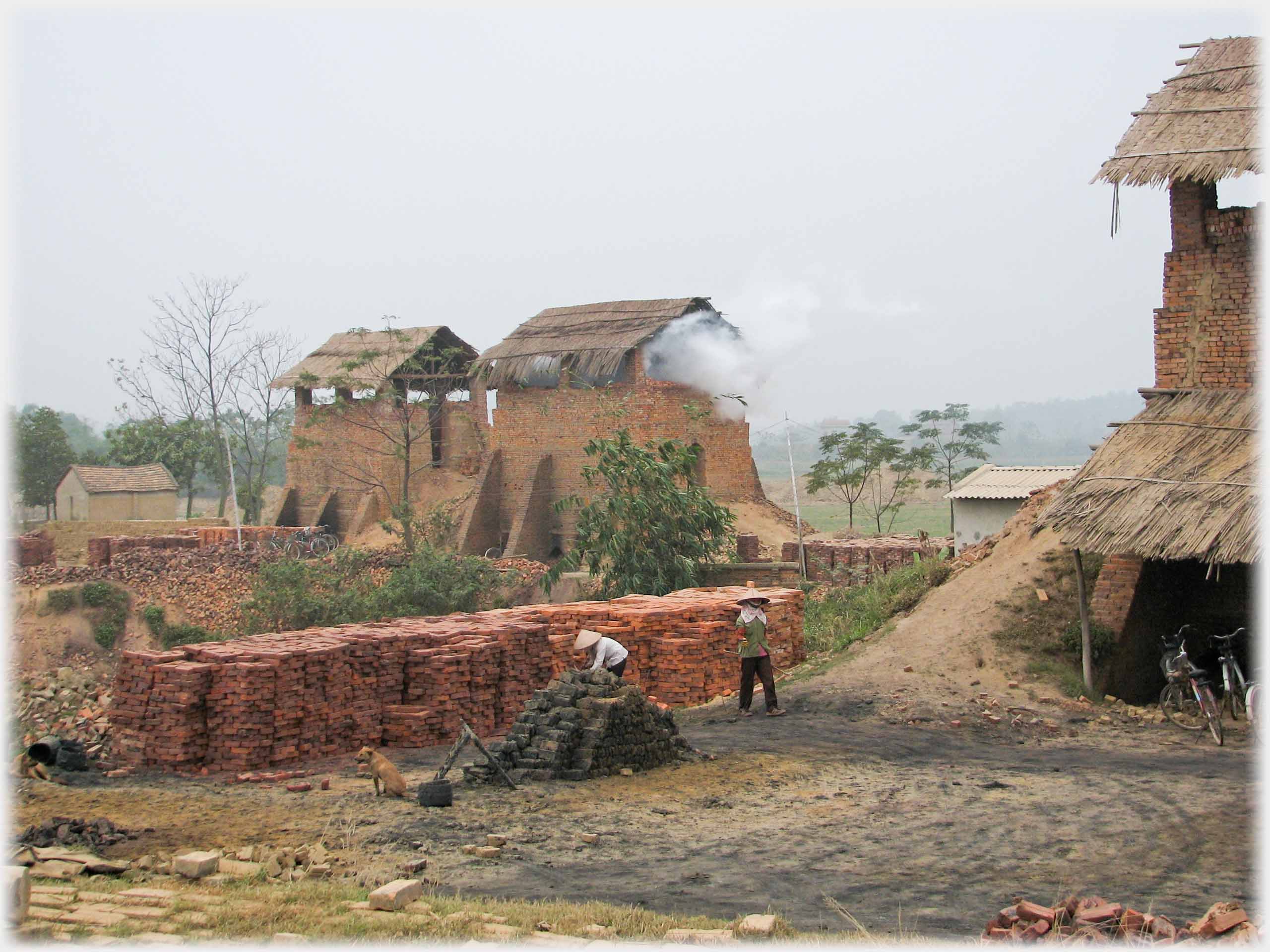 Brick works with thatched roofs, piles of bricks and workers.